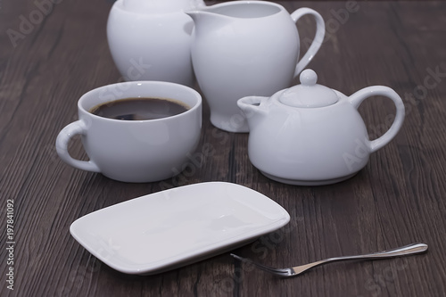 A white cup of coffee and other white utensils on brown wooden table. Breakfast.
