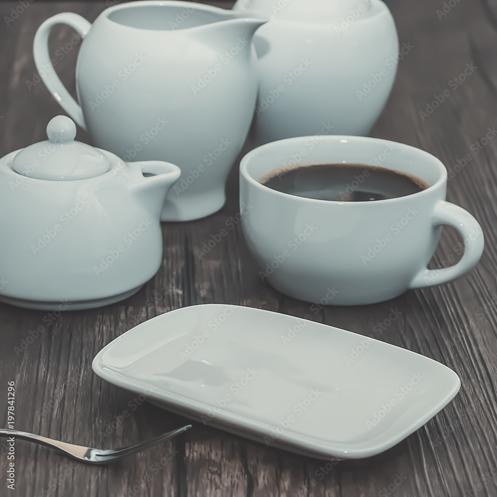 A white cup of coffee and other white utensils on brown wooden table. Breakfast. A white cup of coffee and other white utensils on brown wooden table. Breakfast.