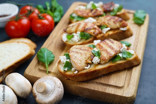 Tasty Italian antipasti bruschetta with grilled chicken, ricotta and basil on wooden board  and cooking ingredients, close up
