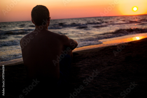 man looks at the sunset on the beach. silhouette of a man who sits on the sand near the sea, in the rays of the setting sun near the sea. place under the text, relax on the sea