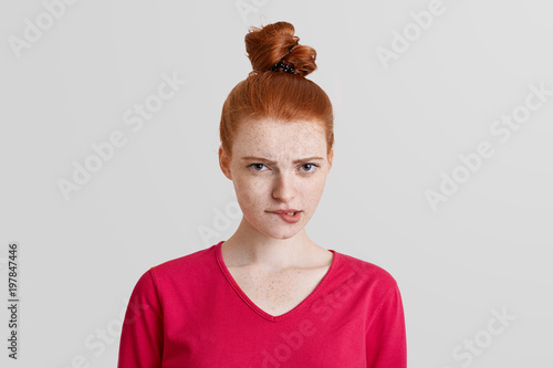 Shot of disconetent redhaired female with hair knot, curves and bites lips, looks with unhappy expression directly into camera, isolated over white background. People, dissatisfaction concept photo