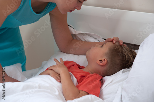 Worried young mother in a blue dress checks the temperature of little cute sick son lying on the bed