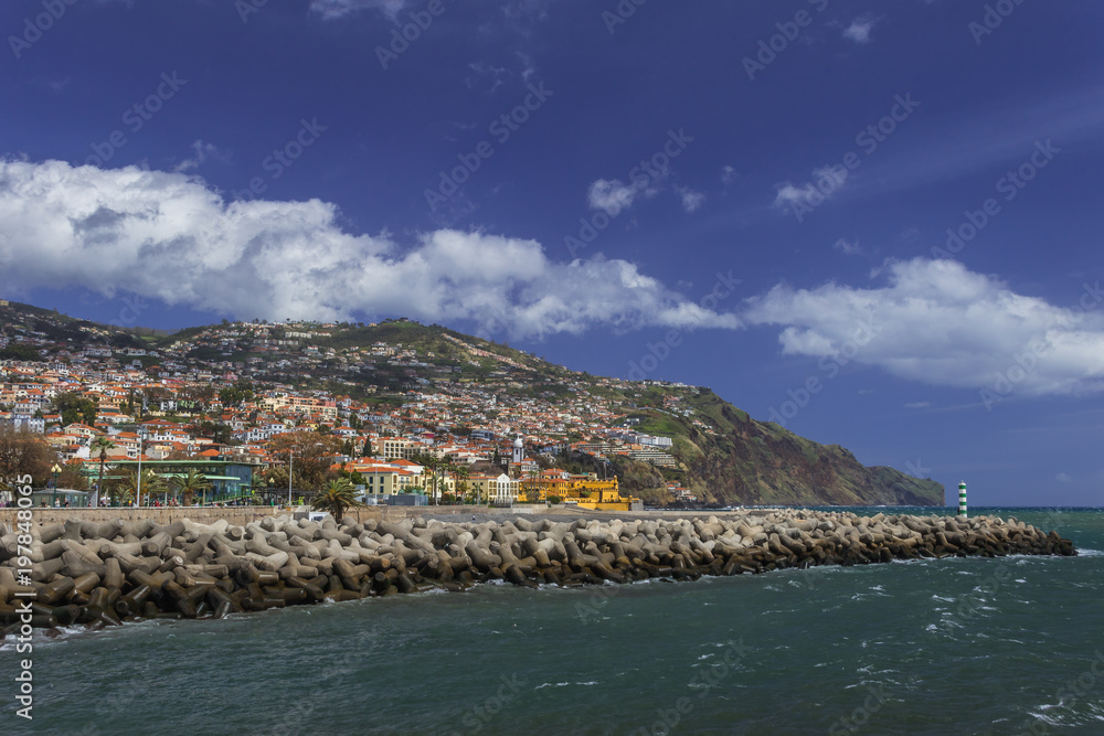 Skyline of Funchal with colorful houses on the hills, beach ath the Atlantic Ocean under the blue sky with fancy white clouds. Vacation on the eternal spring island Madeira