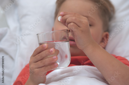 Close-up - Little sick cute cboy holding a pill and a glass of water lying on the bed