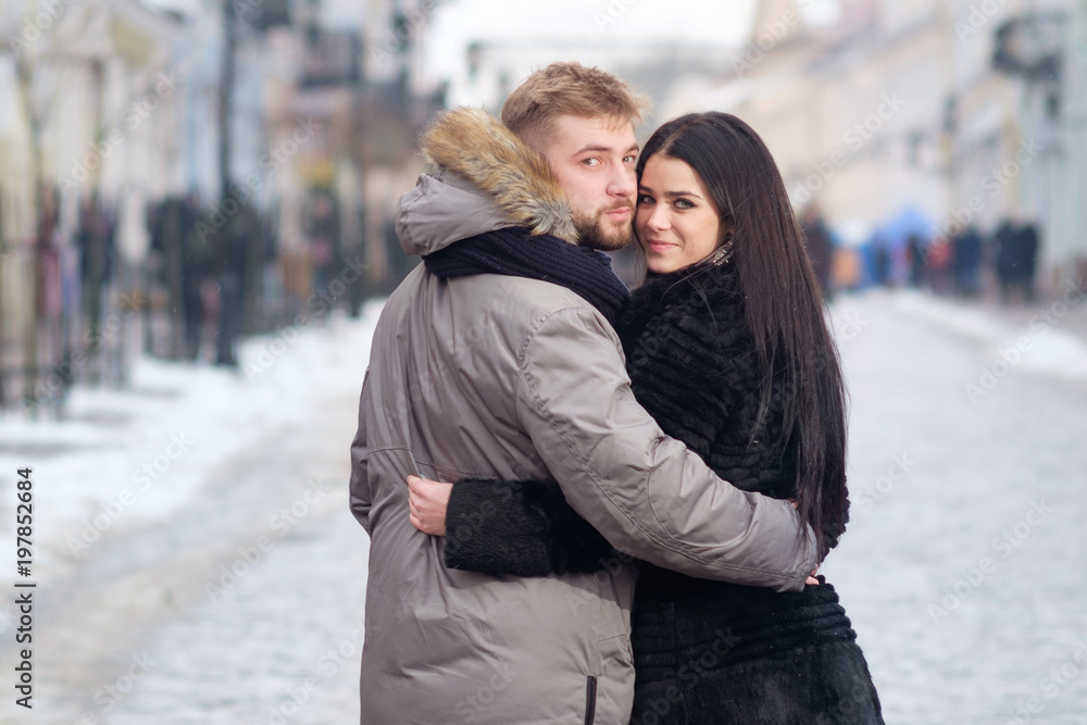 Fototapeta premium young teen couple hug each other standing in center of street uner red umbrella in winter under snowfall, looking in eyes to each other and smiling