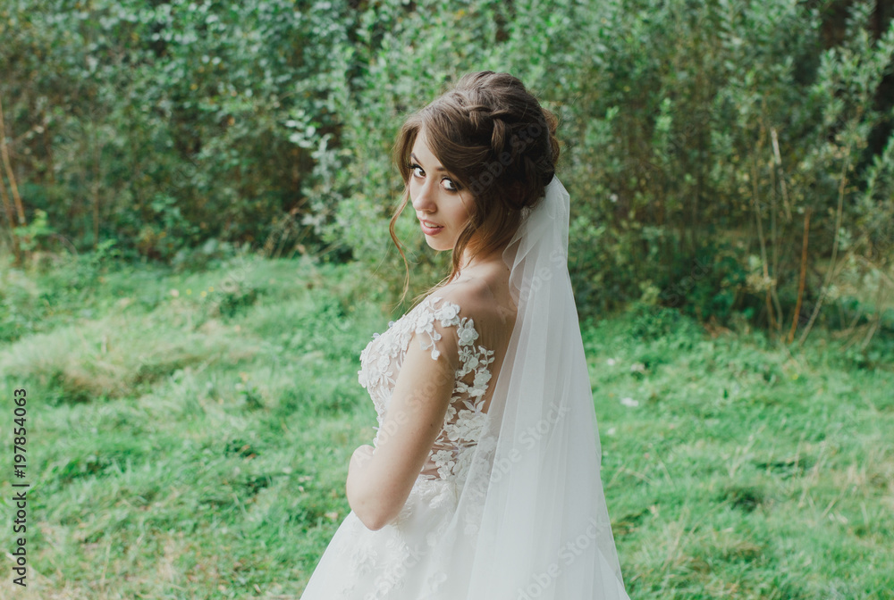 Beautiful bride with elegant simple hairdo is standing in the forest. Low open back white dress with flowers and tulle veil. Stylish hairstyle in outdoors romantic wedding photo. Bridal portrait.