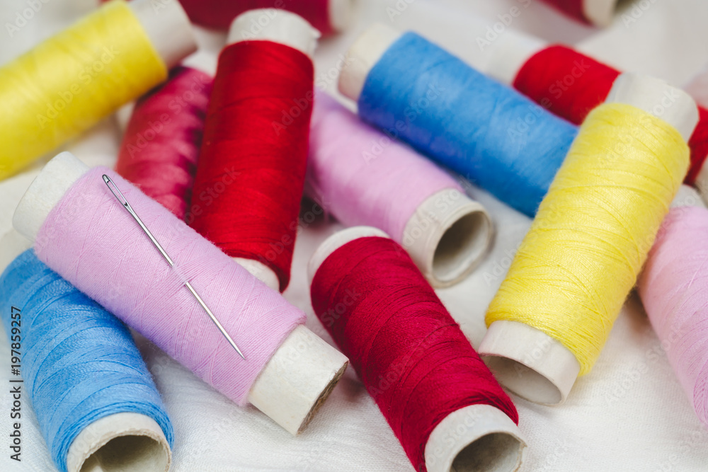 Spools of pink, yellow, blue and red threads and sewing needle on white cotton cloth Spools of pink, yellow, blue and red threads and sewing needle on white cotton cloth