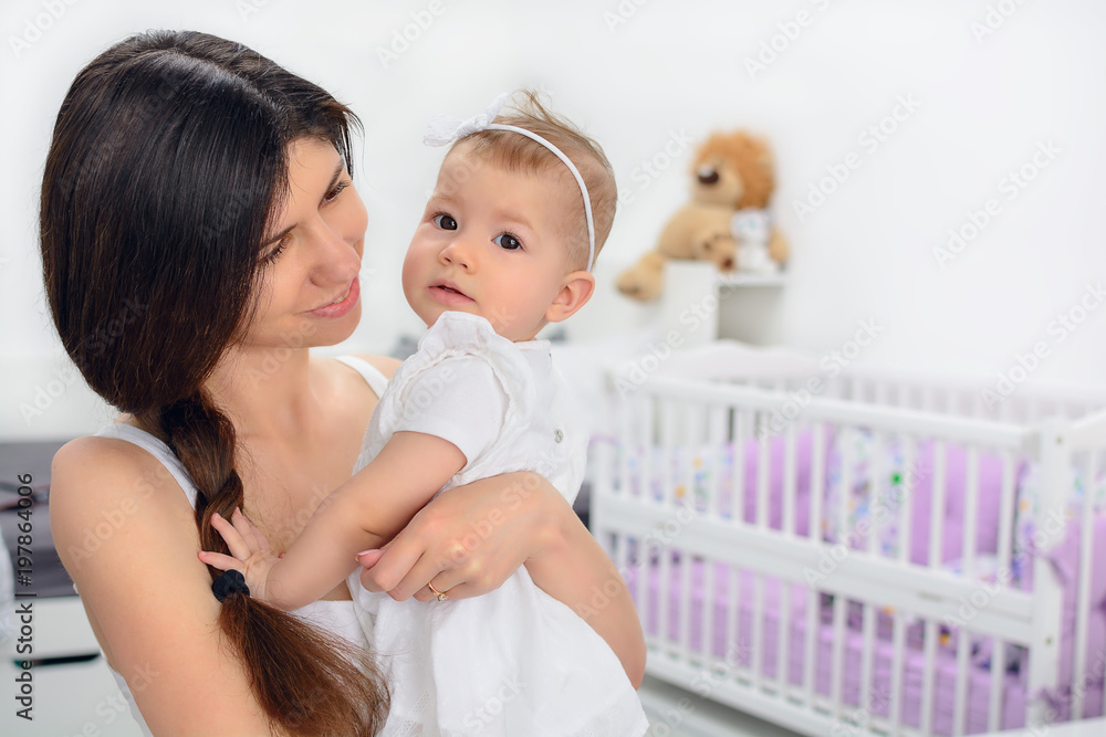 Young mother smiling and holding her daughter child. Woman and baby girl feeling happy in a white bedroom with toys and crib. Nursery interior. Family at home