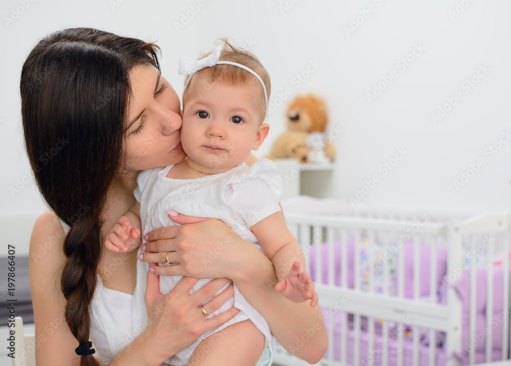 Young mother kissing and holding her daughter child. Woman and baby girl feeling happy in a white bedroom with toys and crib. Nursery interior. Family at home