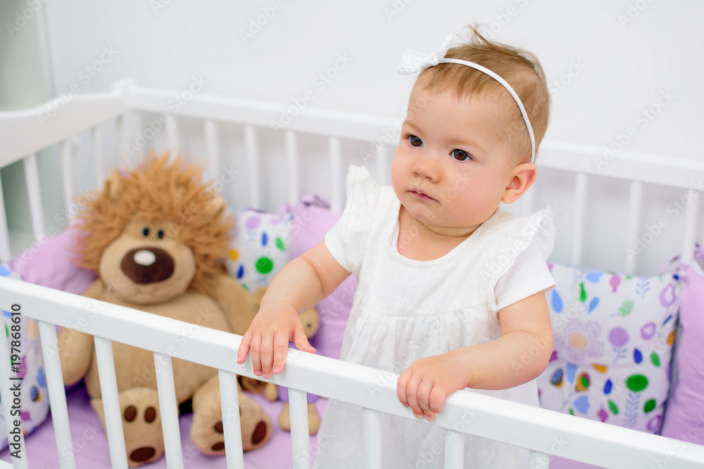 Portrait of 7 month old baby girl standing in white crib in light room