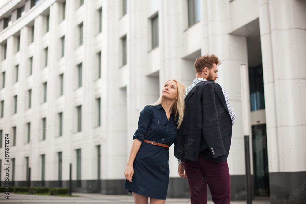 Portrait of young couple standing back to each other in the office clothes, on the background of the building Portrait of young couple standing back to each other in the office clothes, on the background of the building