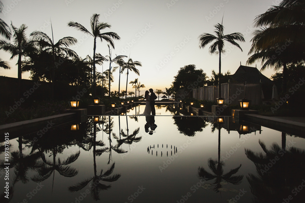 Silhouettes of the bride and groom at sunset. The reflection in the pool