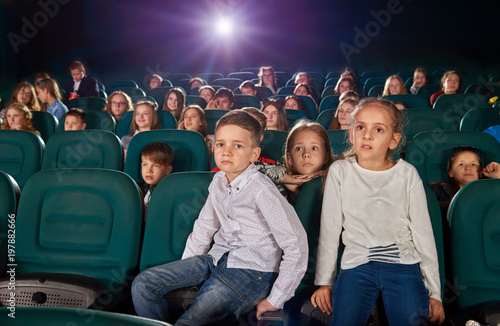 Little boy and girl sitting in the cinema hall by watching new movie or cartoon. Boy is looking at camera, girl is watching film on the cinema screen. Other exited, emotional children on background.