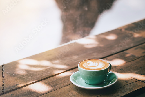 Hot cappuccino coffee in a green cup with latte art on the wooden table in a coffee shop and glass background. Vintage toned.