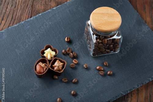 Side view of overturned glass jar with coffee beans and chocolate candies on wooden background, selective focus photo