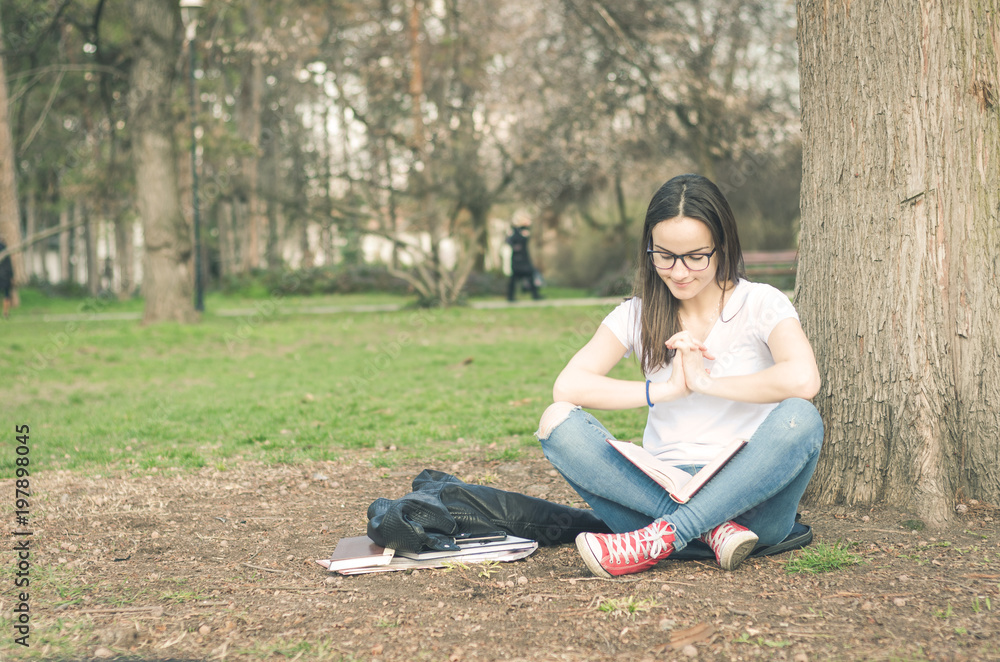 Beautiful young school or college girl with long hair and eye glasses sitting on the ground in the park reading the books and study for exam Beautiful young school or college girl with long hair and eye glasses sitting on the ground in the park reading the books and study for exam