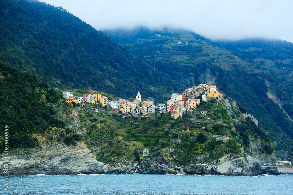 Beautiful scenery of Corniglia viewed from the sea, an amazing village of colorful houses perched on a rocky cliff by the rugged coastline with mountains in background, in Cinque Terre, Italy, Europe