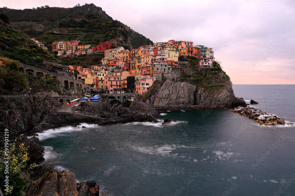 Dusk scenery of Manarola on vertical cliffs by the rocky coast with beautiful sunset reflected on sea water, an amazing village in Cinque Terre National Park, Liguria, Italy, Europe (Long Exposure)