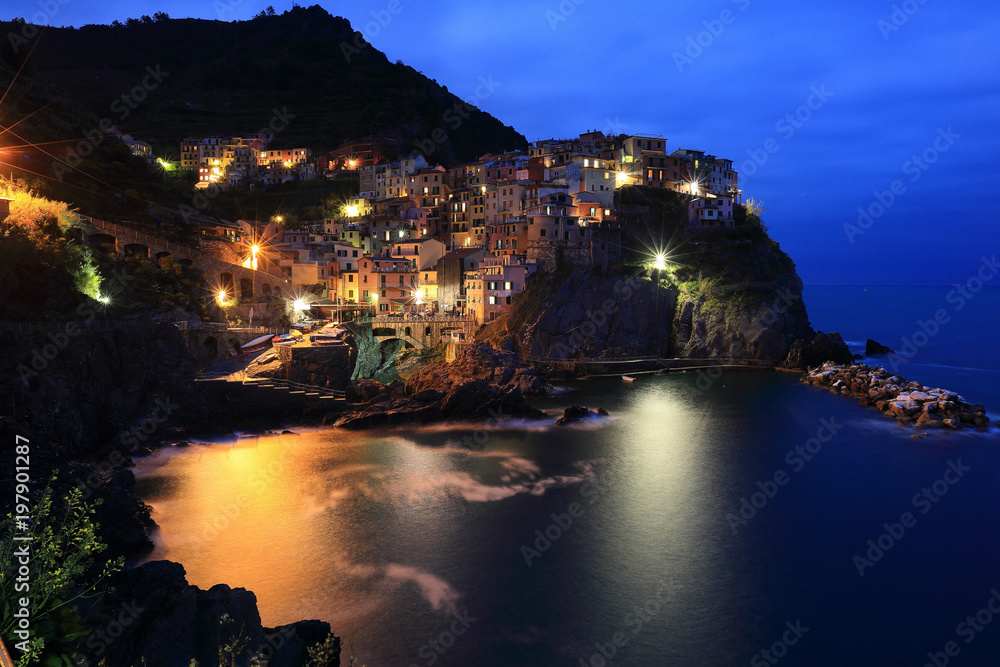 Evening view of Manarola on vertical cliffs by the rocky coast with beautiful lights reflecting on sea water, an amazing village in Cinque Terre National Park, Liguria, Italy, Europe (Long Exposure)