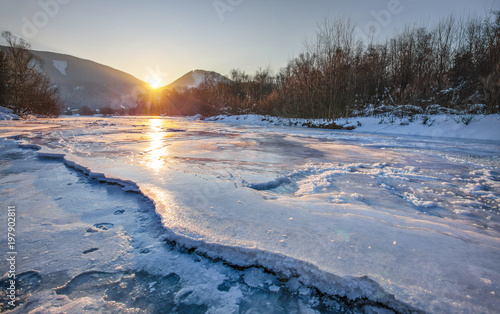 Fototapeta Naklejka Na Ścianę i Meble -  River Bela completely frozen during extreme cold, early morning sun rising behind mountains reflected in ice layer with snow patches. Liptovsky Hradok, Slovakia