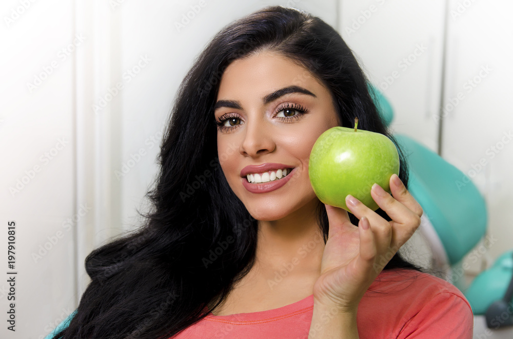 Gorgeous young woman holding green apple next to perfect white teeth , while sitting in dentist chair Gorgeous young woman holding green apple next to perfect white teeth , while sitting in dentist chair