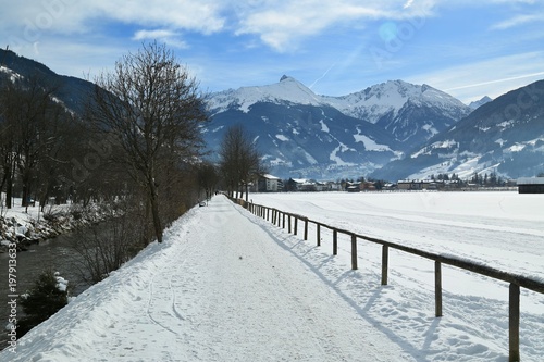 Winter scene with land covered with snow, path along river Gasteiner ache near Bad Hofgastein, Austria, with mountains in distance on a bright sunny day, blue sky, white clouds