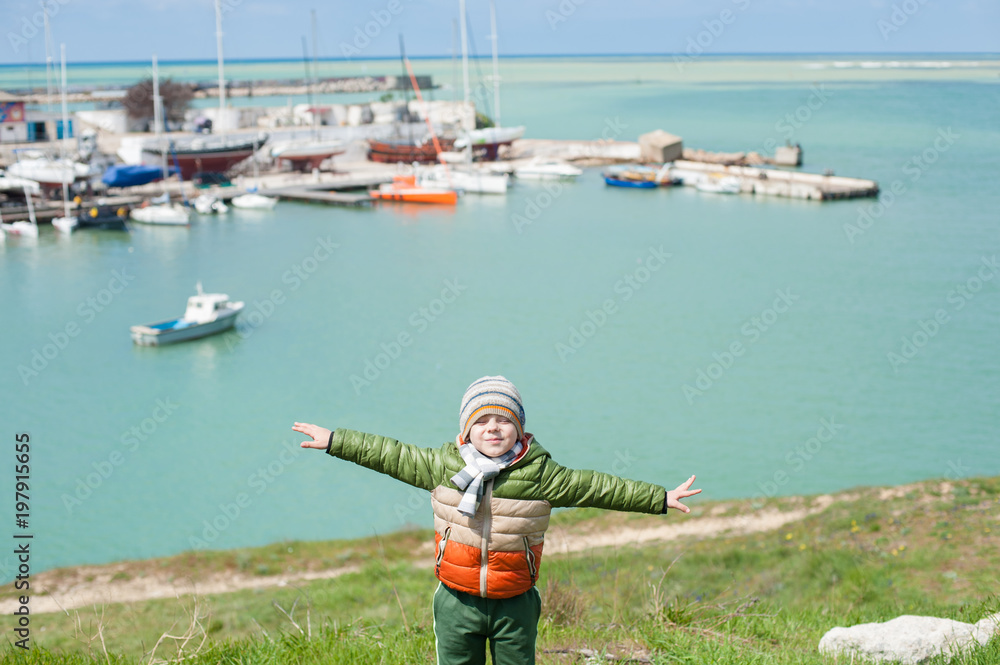 Fototapeta premium dreamy little kid in jacket and hat stands on green hill spreading his hands like wings on background of sea bay with boats in early spring