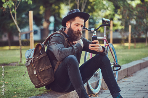 A handsome hipster traveler with a stylish beard and tattoo on his arms dressed in casual clothes and hat with a bag, using the smartphone, sitting on the sidewalk, resting after a bike ride.
