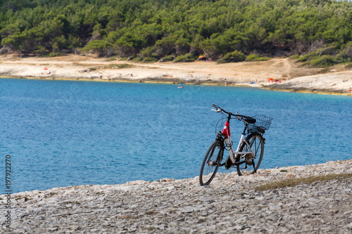 Bicycle stands on stony beach, Kamenjak peninsula by the Adriatic Sea, Premantura, Croatia, swimming people in background, sunny summer day