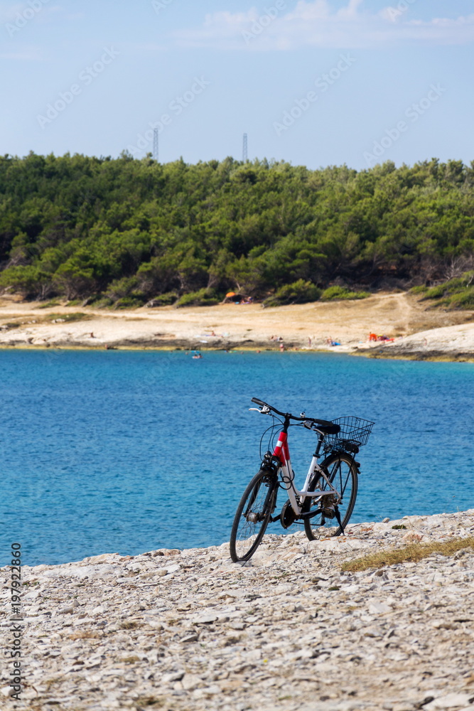 Bicycle stands on stony beach, Kamenjak peninsula by the Adriatic Sea, Premantura, Croatia, swimming people in background, sunny summer day