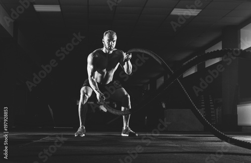 Athletic young man with battle rope doing exercise in the fitness gym. Sports concept. © Dimid