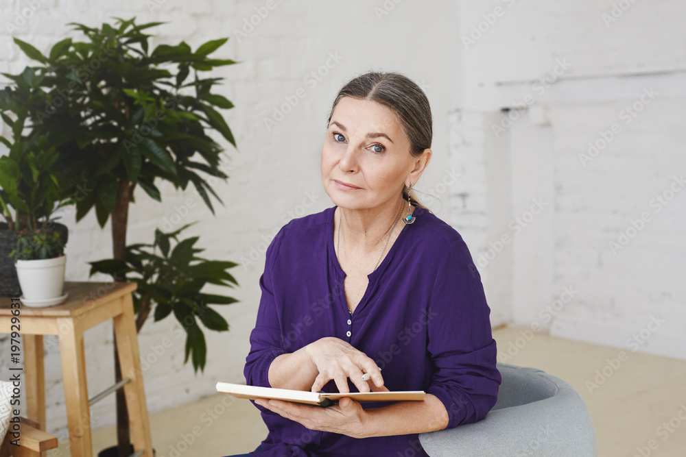 Picture of woman professional psychologist in her fifties waiting for next client, sitting in her modern office on armchair, holding open copybook and looking at camera with serious expression Picture of woman professional psychologist in her fifties waiting for next client, sitting in her modern office on armchair, holding open copybook and looking at camera with serious expression