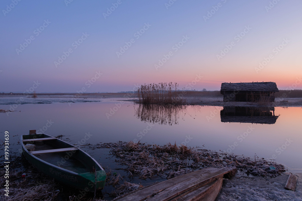 beautiful pastel sky just before sunrise over a wild river surrounded by reeds and an ice-covered shore with colorful boats on them