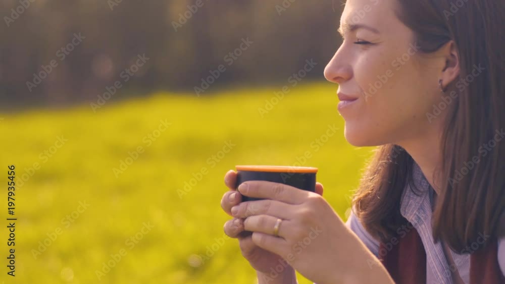 Close-up Side View Young Positive Woman Drinking Coffee From Thermos Cup Sitting On Nature Outside The City In Sunny Warm Weather And Admiring The View. Close-up Side View Young Positive Woman Drinking Coffee From Thermos Cup Sitting On Nature Outside The City In Sunny Warm Weather And Admiring The View.