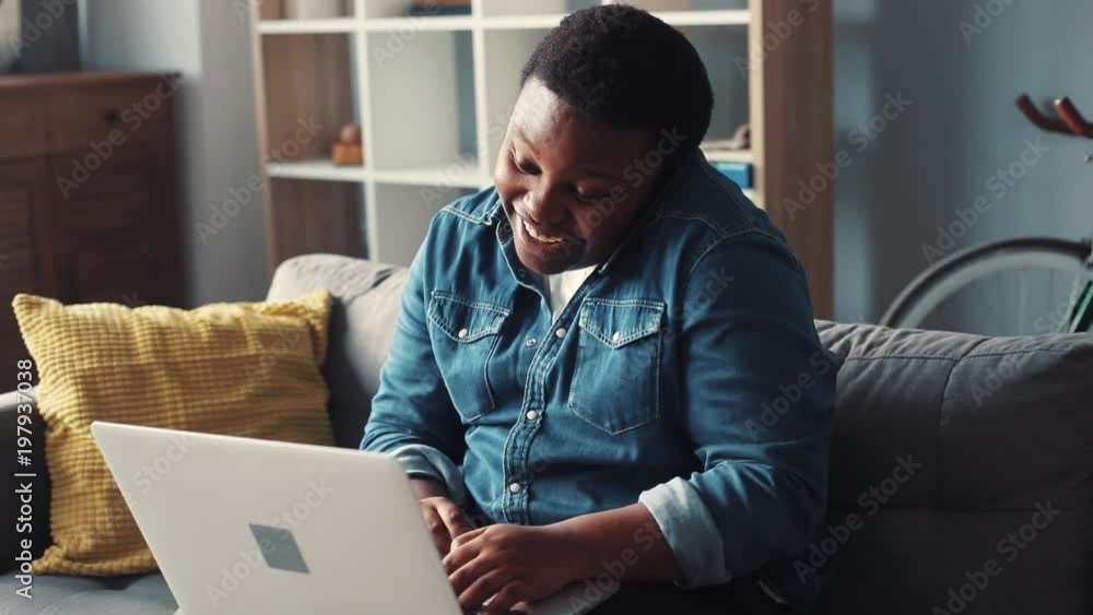 Smile young african american man uses computer home sit on sofa speak on phone programmer looks at monitor screen freelance successful worker businessman engineering Slow motion portrait close up Smile young african american man uses computer home sit on sofa speak on phone programmer looks at monitor screen freelance successful worker businessman engineering Slow motion portrait close up