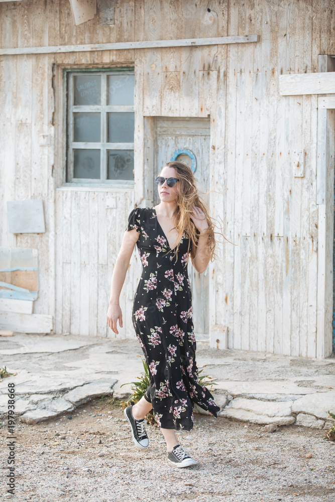 happy and beautiful young woman with sunglasses and flower dress walking in front of a traditional old wooden house in the mediterranean place
