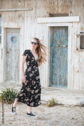 happy and beautiful young woman with sunglasses and flower dress walking in front of a traditional old wooden house in the mediterranean place
