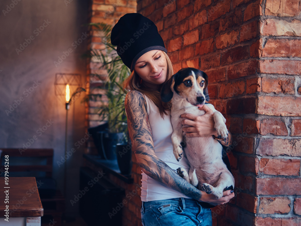 A stylish tattoed blonde female in t-shirt and jeans holds a cute dog. A stylish tattoed blonde female in t-shirt and jeans holds a cute dog.
