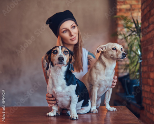 A stylish tattoed blonde female in t-shirt and jeans embraces two cute dogs. © Fxquadro