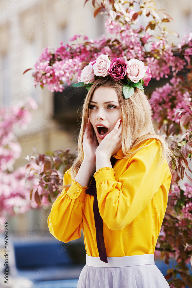 Fototapeta premium Outdoor portrait of young beautiful surprising girl looking aside, posing in street with spring blooming trees. Model expressing joy and excitement.