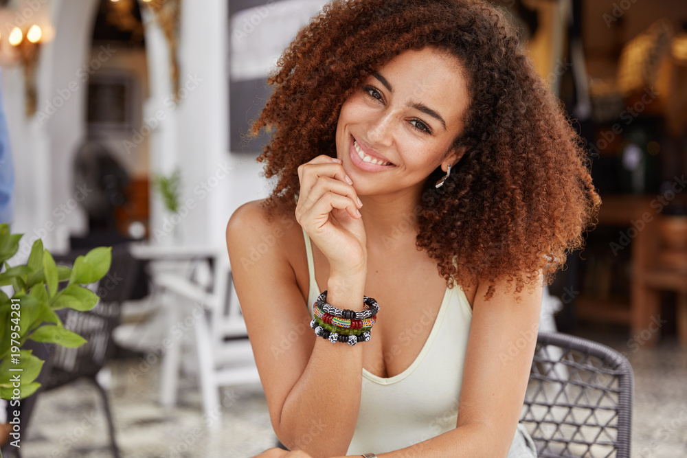 Mixed race dark skinned African American female with bushy hairrstyle, wears bracelet and casual t shirt, being in good mood, has rest in cafeteria. People, positive emotions and rest concept