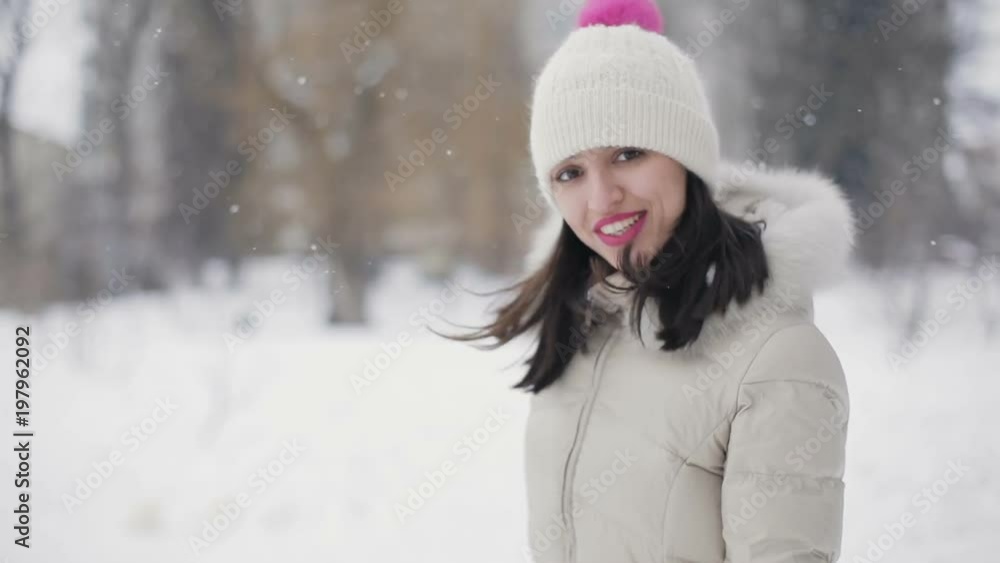 Brunette hair young women with pink lips in beige down coat and hat running away from camera, flirting in snowy central park, snowfall. Slow motion. Brunette hair young women with pink lips in beige down coat and hat running away from camera, flirting in snowy central park, snowfall. Slow motion.