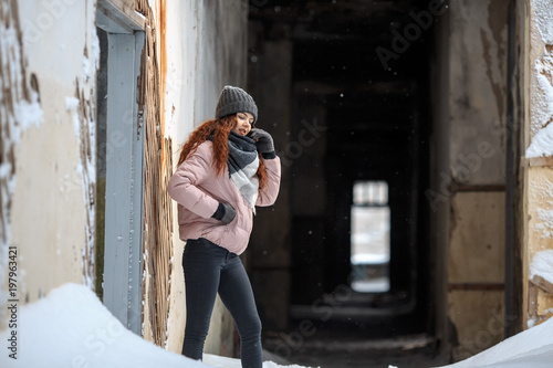 A young cute white girl in a hat warm scarf and gloves is standing in the corridor of a ruined house in winter. Destroyed building. © aleksandr_yu