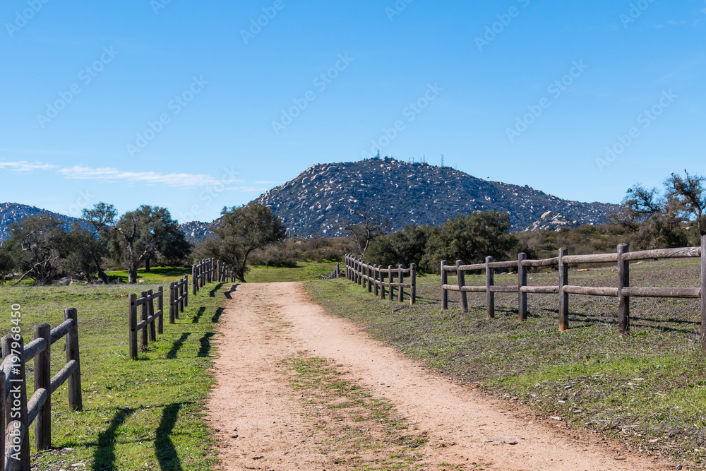 A dirt road lined with a wooden fence leading to Mount Woodson at Ramona Grasslands Preserve in San Diego, California.