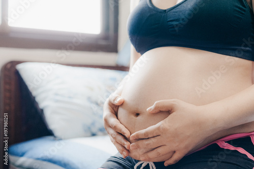 Happy pregnant woman sitting on bed and making heart gesture over window lights background at home, pregnancy, rest, people and expectation concepts