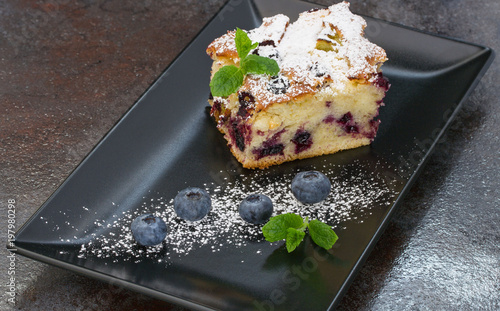 A piece of blueberry pie sprinkled with powdered sugar with blueberries and mint leaves on a black rectangular plate.