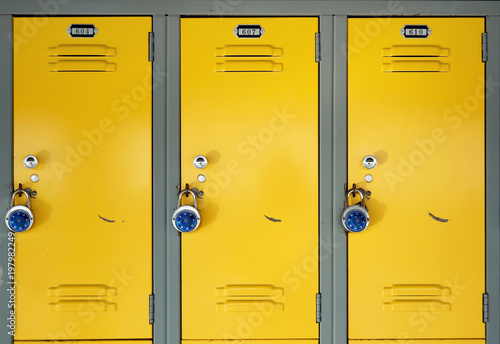 Obraz na plátne Yellow school lockers with locks - Three locker doors of three yellow lockers in the hallway of a school, each with number plates, and a stainless steel combination lock with a blue knob on the face