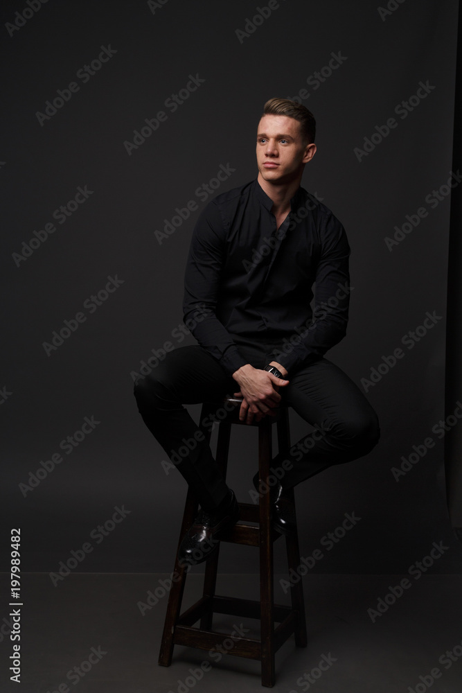 A white man sits on a dark chair in a black shirt and black mills, on a gray background