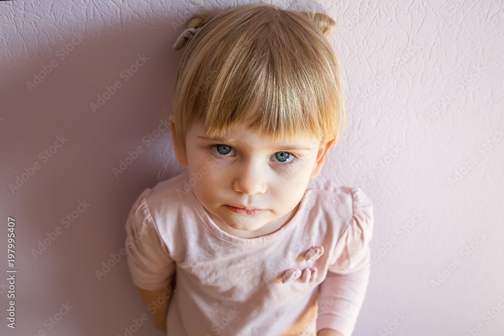 Sad little girl with blond hair is leaning against the wall in a sunny room at home. She looks at the viewer. Top view, close up. Sad little girl with blond hair is leaning against the wall in a sunny room at home. She looks at the viewer. Top view, close up.