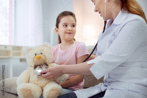 Great patient. Pretty little girl looking at female doctor who sitting next to her and treating plush bear